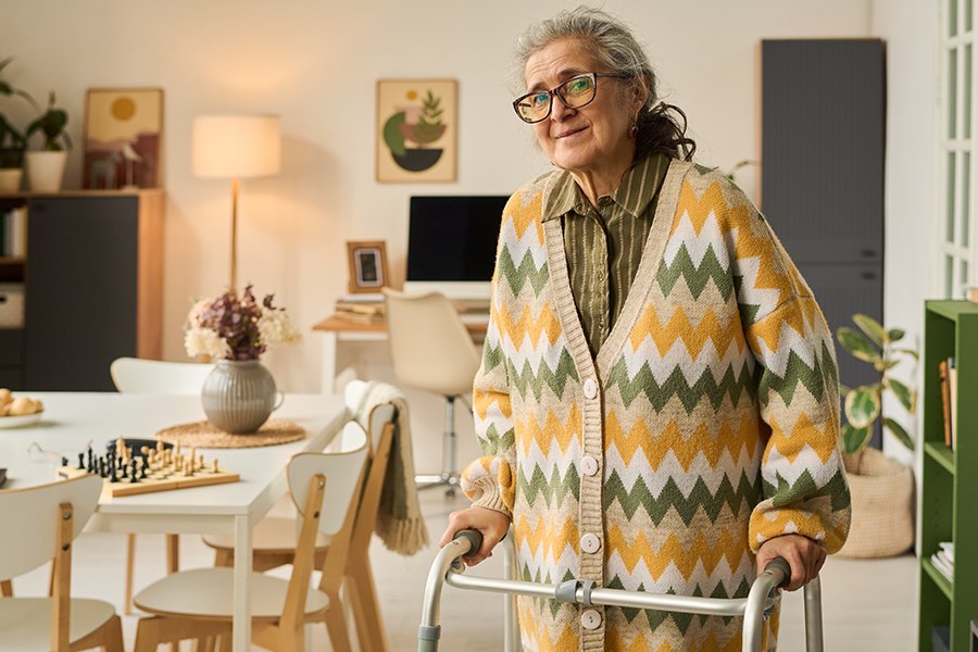 An older woman standing with her walker in her own living room, a chess set on the table behind her