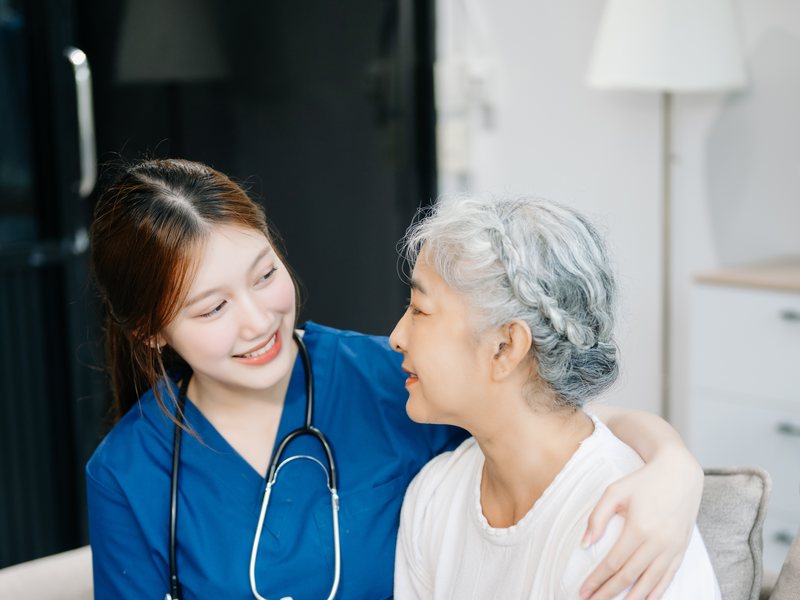A nurse in scrubs sitting with her arm around an older woman at home, the two of them smiling at each other